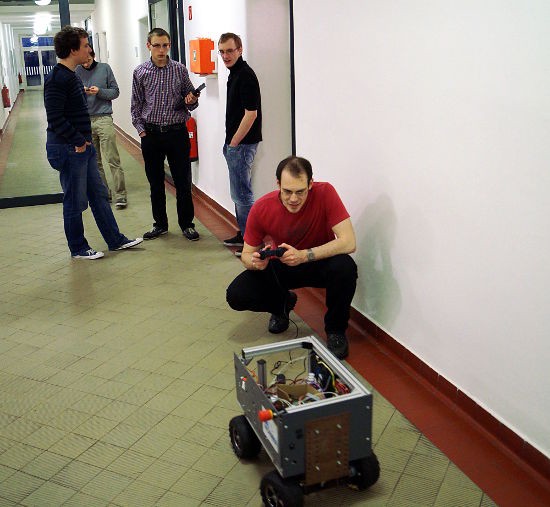 Der Roboter fährt durch den Flur und wird von einem Kamaro mit dem Gamepad gesteuert. Einige weitere Kamaros unterhalten sich im Hintergrund