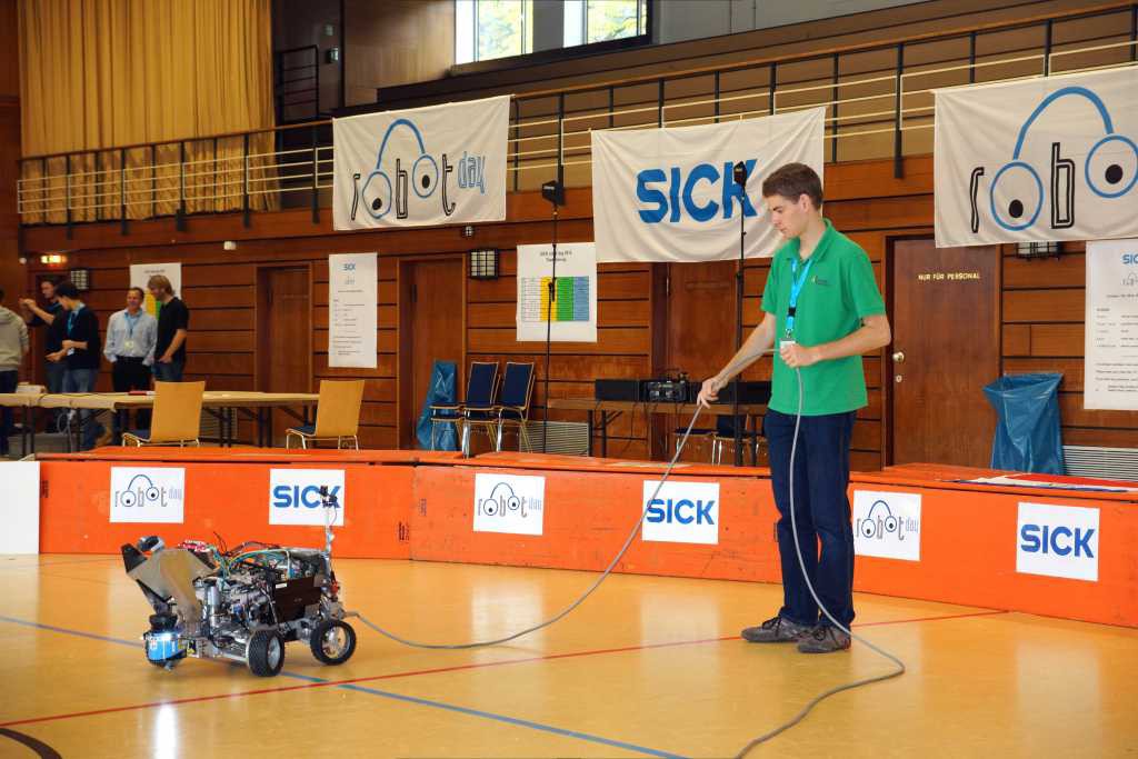 Beteigeuze steht in der Arena des SICK Robot Day 2014 und ist per Kabel mit Strom versorgt. Ein Kamaro steht daneben und hält das Kabel.